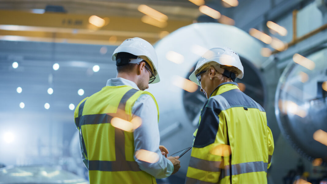 Eine Frau und ein Mann stehen in gelber Sicherheitskleidung und einem Helm vor einer großen Maschine und schauen gemeinsam auf ein Dokument