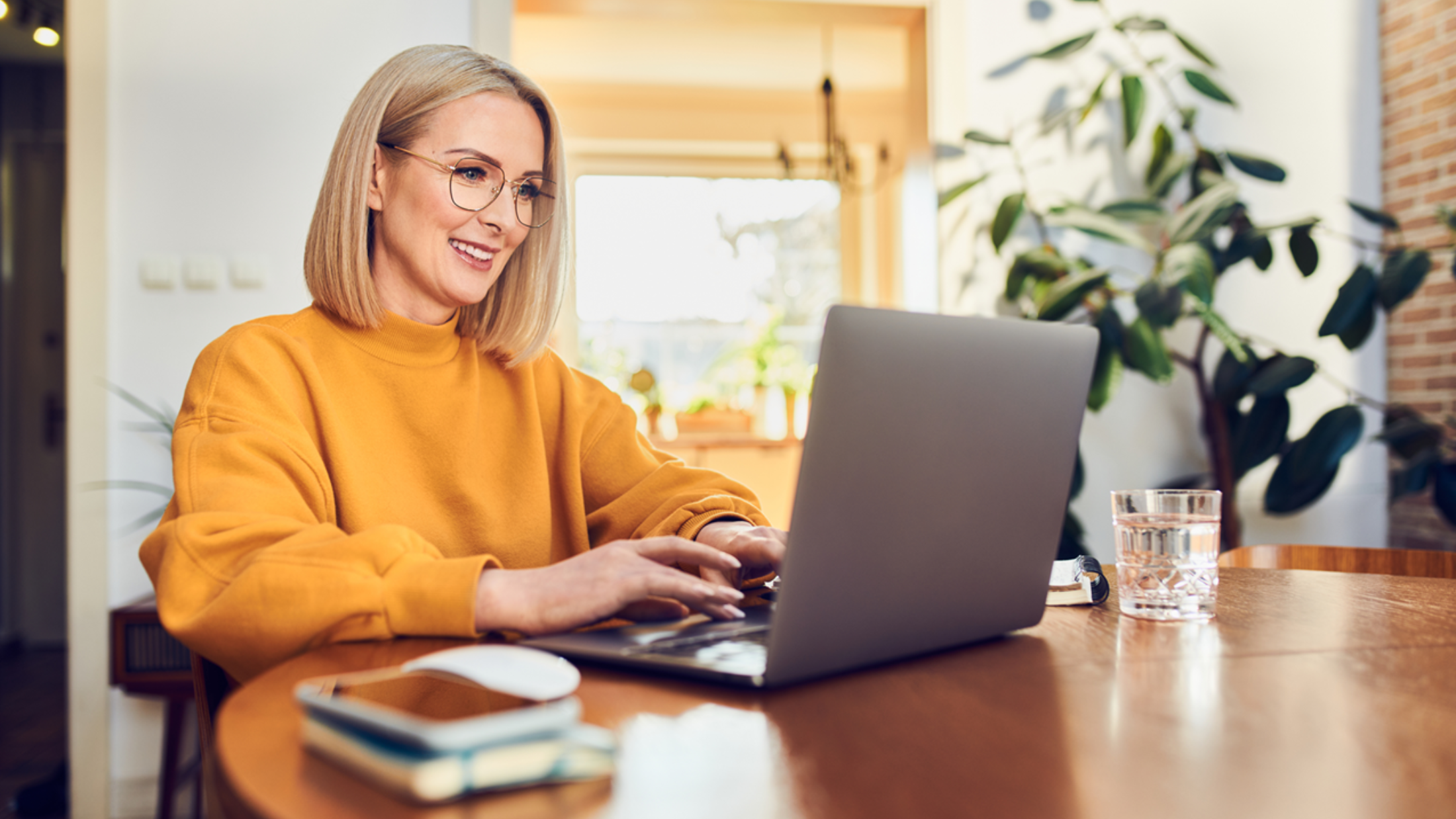 Eine blonde Frau mit Brille und gelbem Pullover sitzt an einem runden Tisch und arbeitet an ihrem Laptop.