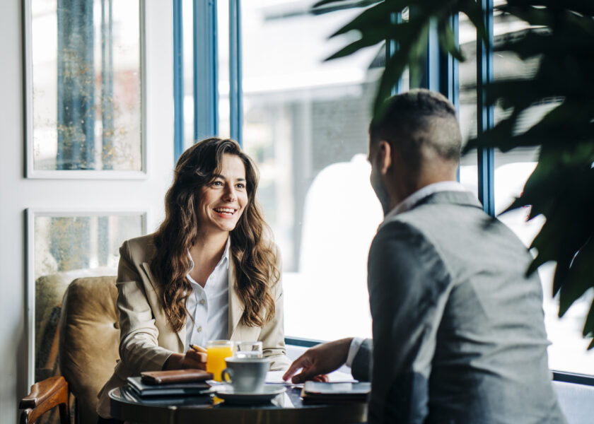 Zwei Personen sitzen in einem Café an einem kleinen Tisch und führen ein Gespräch, während Getränke und Unterlagen vor ihnen liegen.