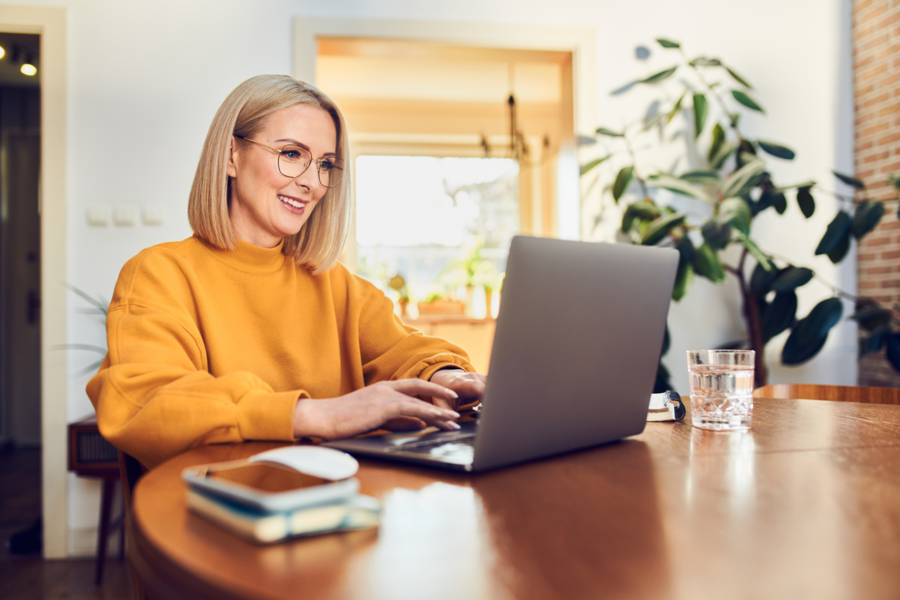 Eine blonde Frau mit Brille und gelbem Pullover sitzt an einem runden Tisch und arbeitet an ihrem Laptop.