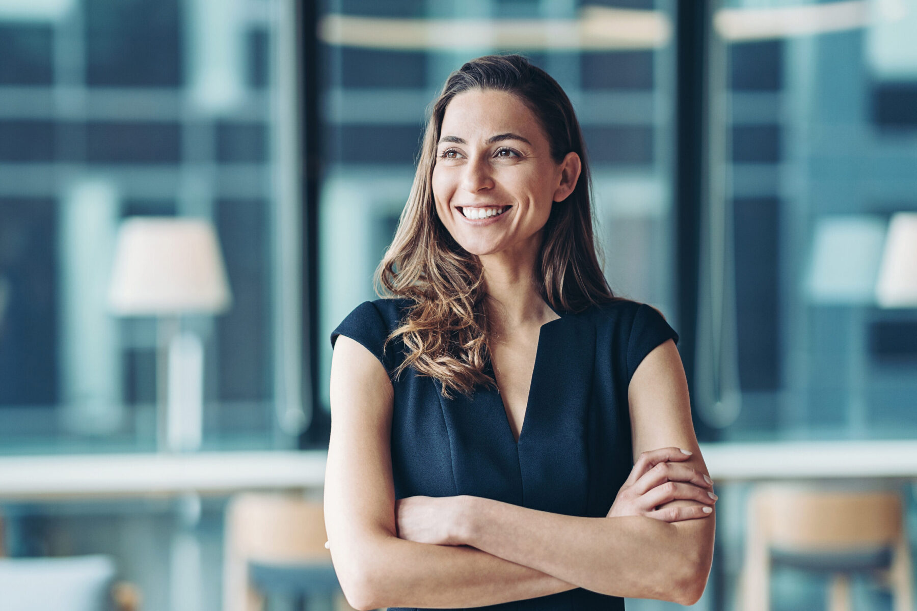 Frau in Businesskleidung, die im Büro steht, ihre Hände verschränkt und selbstbewusst in lächelt