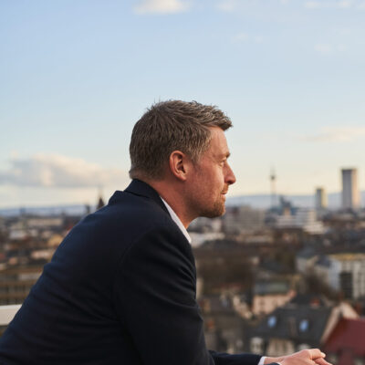 Ein Mann in einem schwarze Sakko lehnt an einem Balkongeländer und blickt auf die Skyline einer Stadt.